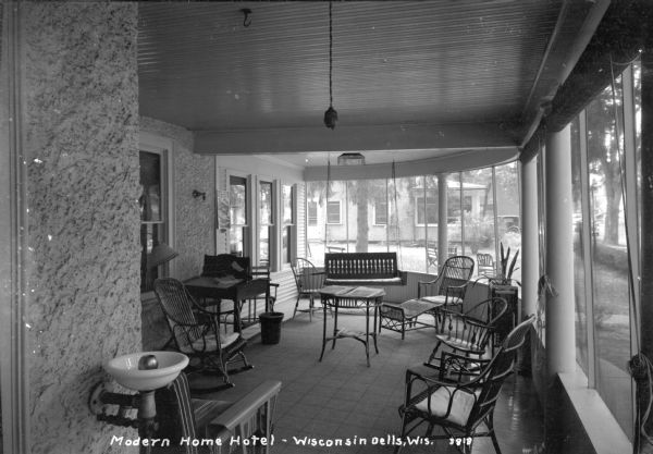 Front porch at Modern Home Hotel. There is a drinking fountain in the foreground on the left. The furniture on the porch includes a desk, rocking chairs, a porch swing, a lounge chair, and a table with magazines and newspapers. A poster advertising an upcoming dance band is posted above the desk. Chairs and benches are on the lawn outside.