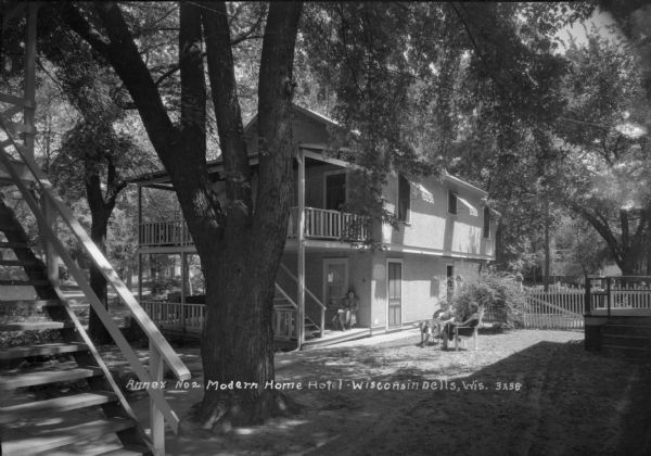 View of an annex with more guest rooms, and exterior stairs that rist from the porch up to a balcony. Women are sitting on the porch and the lawn. On the left in the foreground is a set of stairs leading up to a balcony of another building.