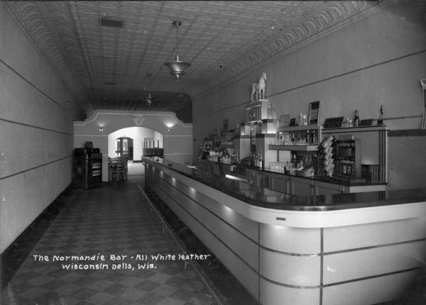 Interior view of a tavern with an all white leather bar, and a stamped tin ceiling. Glasses, bottles of liquor and bags of chips and peanuts are on display on the wall behind the bar, including ceramic figurines of white horses which have "White Horse Whiskey" written at the base. On the other end of the bar is a jukebox in the corner, next to an archway leading to another room with tables and chairs.