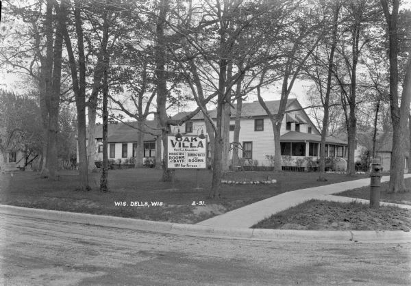 Three-quarter view from road of a guest house with a large porch. A large sign on the lawn in front advertises running water in the rooms and a rate of $1 per person. A lawn swing and benches are on the lawn. There is a fire hydrant on the corner next to the sidewalk.