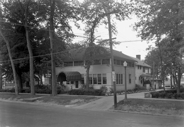 Exterior view of the Olson's Rooms hotel, with an arched awning over the entrance, and a driveway on the right. There are benches near a stone building on the left behind trees. In the background on the left a man stands in front of a store window, which appears to have a painted sign reading: "Parson's."