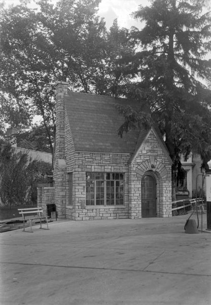 A small stone building with a steeply pitched roof, chimney, large, paned windows and arched doorway. Benches are on the left and right of the building. On the right is a gas pump. The Olson's Rooms hotel building is in the background.