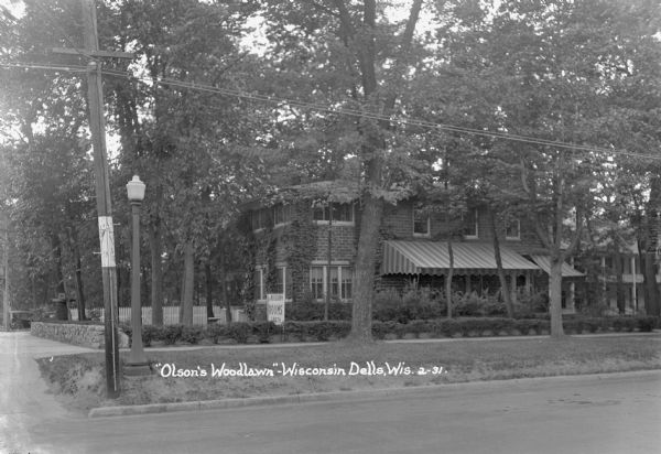View from street of Olson's Woodlawn Rooms, a large brick building with striped awnings over some of the windows on the first floor. A low stone wall is on the left, and a picket fence is in the back. Another building with a porch and a balcony is in the background on the right.