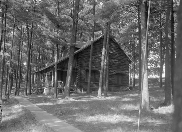 View down sidewalk towards a log building among the pines. The sidewalk leads to the large front porch with steps in the front and on the side.