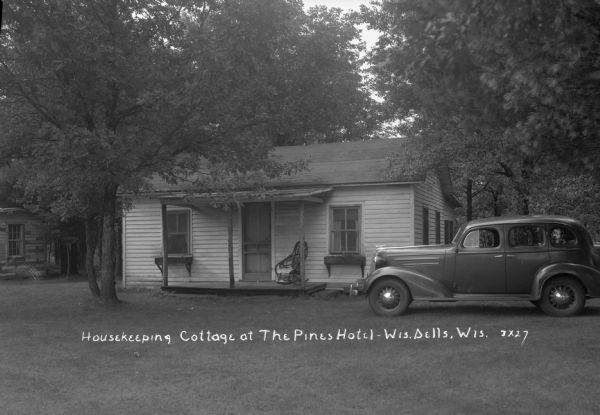 Small cottage with a rocking chair on the porch. An automobile is parked in the lawn on the right, and another cottage is on the left.