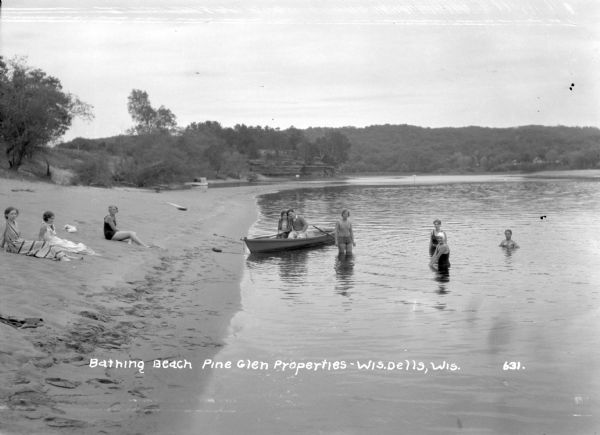 View along shoreline of a bathing beach on the Wisconsin River at Pine Glen Properties. Bathers on sitting on the beach on the left. Four more people are standing in the water, and a couple poses in a rowboat.