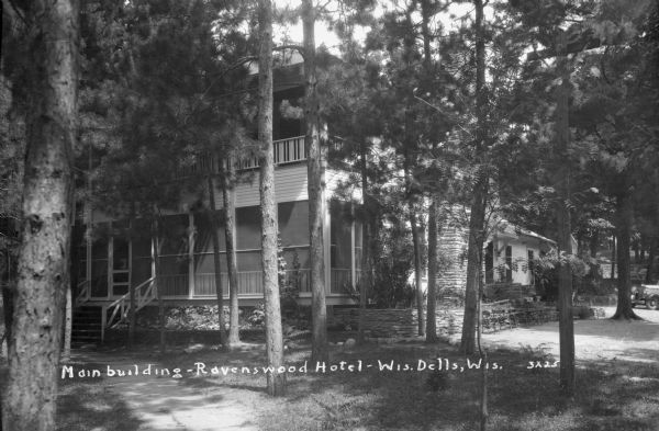 Exterior view of the Ravenswood Hotel set amongst the pines, with a large screened porch and balcony above. Benches are along a stone wall in front of a stone chimney. An automobile is parked in the background on the drive on the right.