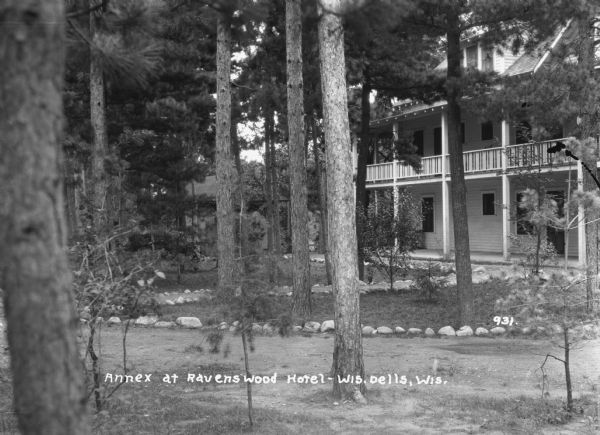 View across stone-lined paths toward the annex, a three-story building with a porch and a balcony above. Pines trees are on the grounds, and there is a smaller building behind the trees on the left. The Wisconsin River is below in the far background.