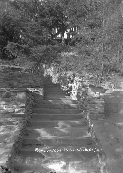 View looking up the stone steps leading up to the Ravenswood Hotel, obscured by trees at the top of the hill. Two women are sitting on the stone wall on the right.