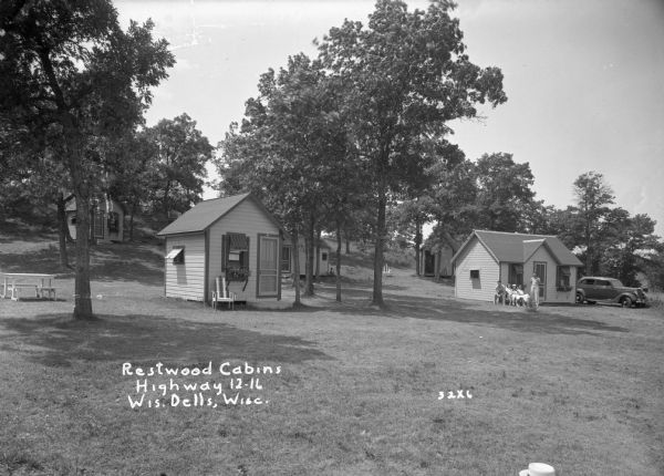 Group of small cabins on a slope, with a picnic table on the left, and an automobile on the right. A group of three women and two men posed in front of the cabin on the lower right.
