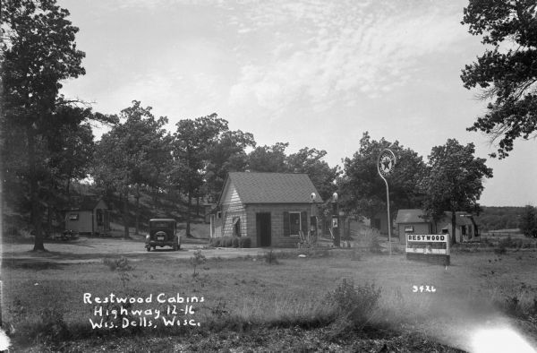 Texaco station, with an automobile parked next to the a stone building which has two pumps, at Restwood Cabins. Cabins surround the station, and there is a low hill in the background.