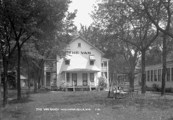 View across lawn of the annex of Hotel Van. There are two entrances and two exterior stairs on either side of the building. Two women are sitting in the lawn swing on the front lawn. Another woman stands on the left balcony looking down at a dog on the landing of the steps.