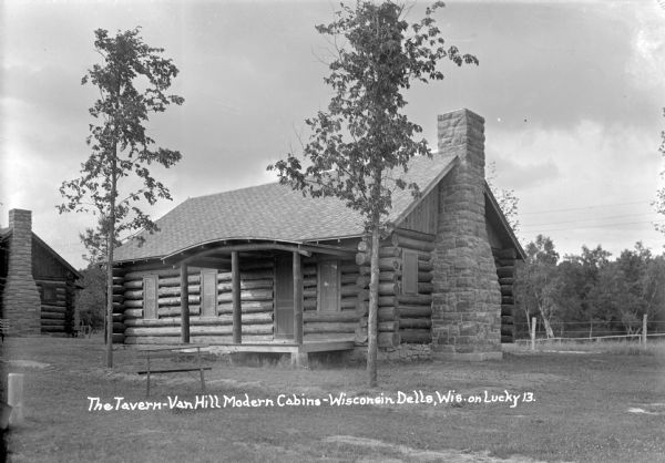 Exterior view of a guest cabin called "The Tavern" at Van Hill Modern Cabins. A stone chimney is on the right, and the open porch has an arched roof and log columns. Young trees frame the entrance, and a bench sits on the lawn in front. Another cabin is on the left in the background.