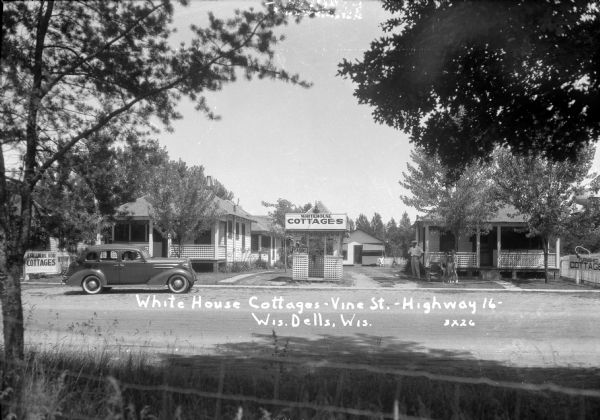 View across road of the cottages at the White House Hotel, with an information kiosk along the sidewalk between them. A group of people are in front of the cottage on the right. An automobile with passengers is parked at the curb.