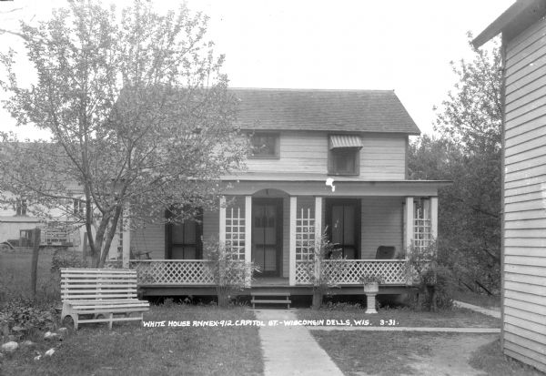 View of the Annex of the White House Hotel. There are three entrances on the porch. There is a porch swing on the left, and a bench is in the yard in front. In the background a truck is parked in front of a large building.
