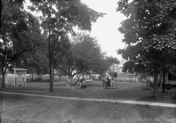 View across sidewalk of the courtyard of the White House Hotel. The Annex is on the right. A gazebo is on the left, there are benches and a lawn swing in the center. The property is surrounded by white picket fences. Several people and a dog are gathered on the lawn.