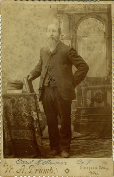 Full-length cabinet card portrait in front of a painted backdrop of Carl Nielson [as written on published Roster of Wisconsin Volunteers, Volume 1 and on the Regimental Descriptive Rolls, Volume 20 ; written as Carl Nelson on card backing of portrait], a private in Company F, Wisconsin 15th Infantry. The following information was obtained from the Regimental and Descriptive Rolls, Volume 20: He resided in Door County, Wisconsin. On February 2, 1862, he enlisted in Door County, Wisconsin and was mustered into service in Madison, Wisconsin on March 1, 1862, at the age of 32. He was in a hospital in Iuka, Mississippi starting August 20, 1862 and was eventually discharged on April 10, 1863 at Quincy, Illinois for disability.