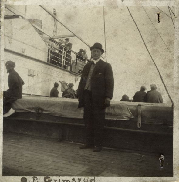Full-length portrait of Olaus Pederson Grimsrud standing on the deck of a ship. Other people are sitting and standing behind him.