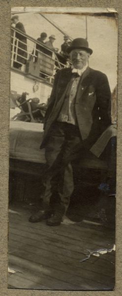 Full-length portrait of Olaus Pederson Grimsrud standing on the deck of a ship. Other people are in the background.
