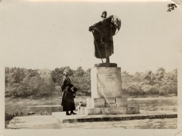 Older woman standing outdoors next to a sculpture of Thorfinn Karlsefni, an Icelandic-born Scandinavian explorer of North America. Behind her is the Schuylkill River and the tree-lined opposite shoreline.