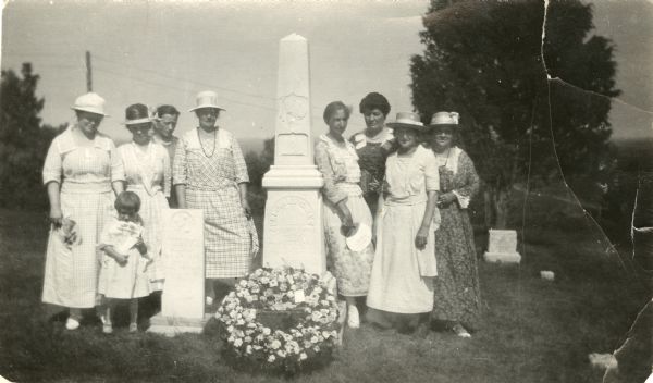 Outdoor group portrait of women and one child alongside the graves of Colonel Hans Heg, commander of the 15th Wisconsin Infantry, and his daughter Annetta. There is a large wreath of flowers at the base of Colonel Hans Heg's grave.