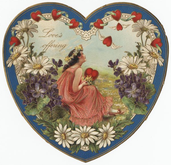 Valentine's Day card in the shape of a heart, with a woman in a long pink dress sitting in a meadow as the central image. She is holding a heart and flowers and has a garland of flowers in her hair. The heart has a blue, metallic gold, and white border, with hearts above and flowers below. Two of the hearts have wings. The text "Love's Offering" is in the upper left corner. Chromolithograph, embossed and die cut.