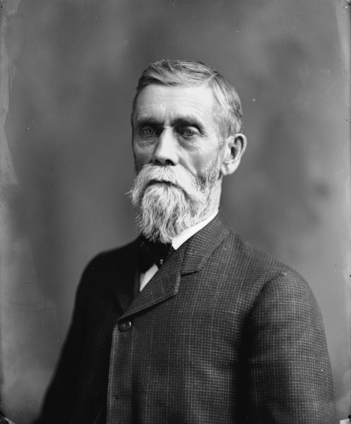 Waist-up studio portrait of E. Rutledge. He has white hair, a moustache and beard, and is wearing a plaid suit and cravat.