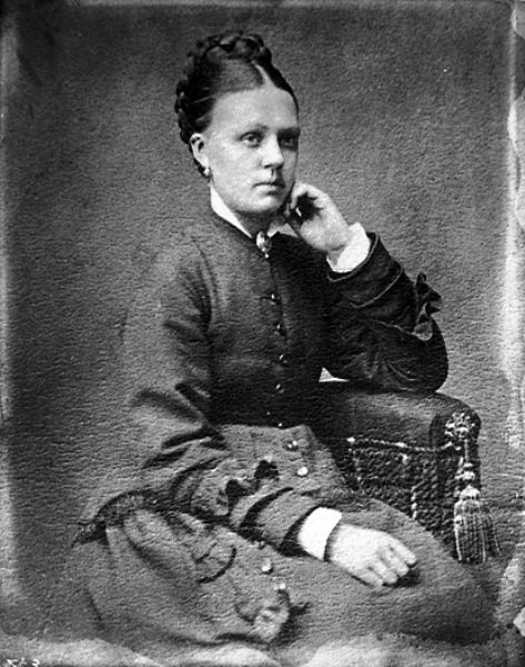 Studio portrait of a woman sitting in a chair. She is wearing the dress fashionable around 1870.