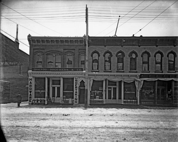 Hoenig Brothers & Co. two-story hardware and undertaking storefront. A man stands to the left of the building.