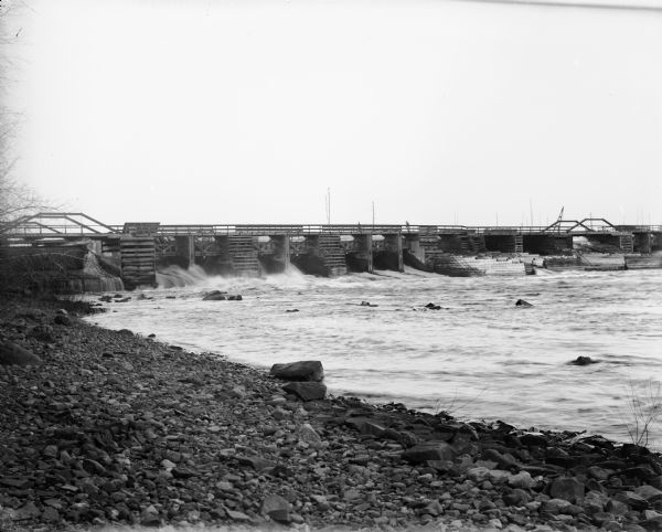The Holcomb[e] or Little Falls Dam on the Chippewa River, with rocky shoreline in foreground.