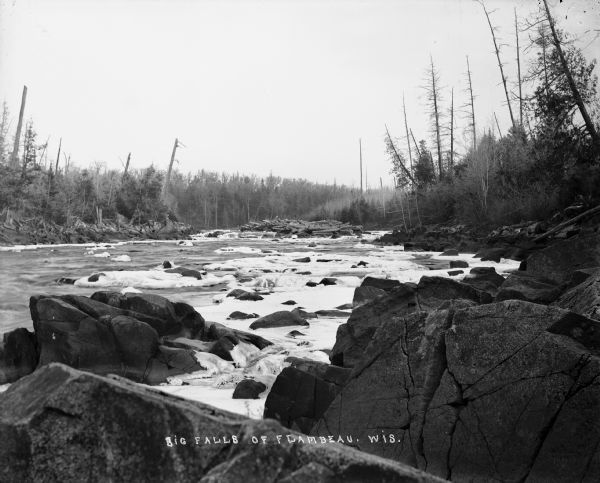 Big Falls, looking down the Flambeau River. There are large boulders in the foreground and patches of ice in the river.