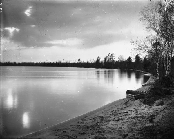 The curved shoreline of Round Lake. A rowboat is drawn up to the sandy shoreline on the right. The surface of the water is smooth.