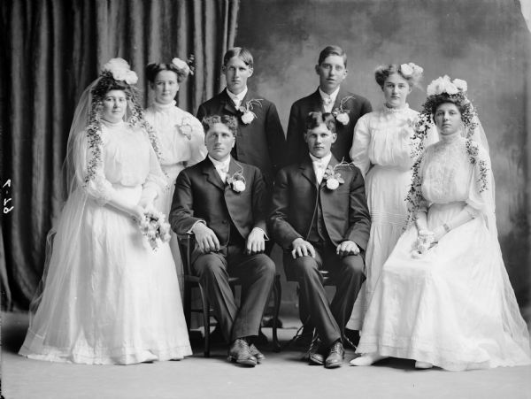 Eight members of the wedding party of Albert Bowe. Four men in dark suits wearing boutonnieres are seated and standing in two rows in the middle of the portraits. Four women in white dresses and elaborate headdresses are seated and standing in two rows to the outside of the portrait.