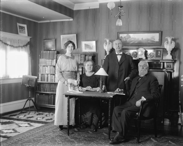 Four members of the Bailey family, two men and two women, stand and sit around a small table in front of a fireplace. There is a music stand below the window, and numerous paintings and/or photographs hanging on the walls. A clock and two vases stand on the mantelpiece, and rugs cover the floor. A bookcase filled with books lines half the wall behind the family. Three of the four members of the family are holding reading material, either books or magazines.