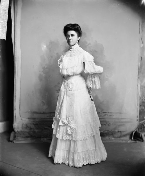 Full-length studio portrait of Mrs. Merkle, wearing a frilly white dress with lace at hem, cuffs, and neckline.
