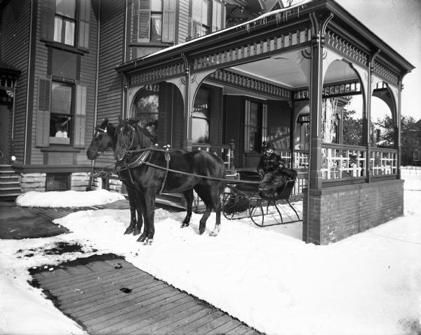 Winter scene with William Irvine (1851-1927) seated in his sleigh next to his daughter Ruth (1891-1987) wearing a hat and wrapped in furs, Chippewa Falls, Wisconsin. Mr. Irvine is holding the reins of two horses wearing bells and hitched to the sleigh. The sleigh stands under a portico attached to Mr. Irvine's house. There is snow on the ground, and on the left a woman can be seen in a first floor window holding the shade.