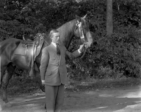 Tom Cummings, wearing a suit and cravat, stands holding the reins of his saddled horse, who is wearing a ribbon reading "1st Premium."