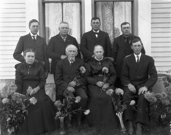 Group portrait of eight members of the Meinen family on the occasion of the golden anniversary of two of its members. The portrait was taken in front of two windows of a house. Two men and two women are seated in the front row and four men stand in the back row. All are dressed in black dresses or suits. The older man and woman seated in the center are holding flowers, and flowers in vases stand on the ground in front of the group.