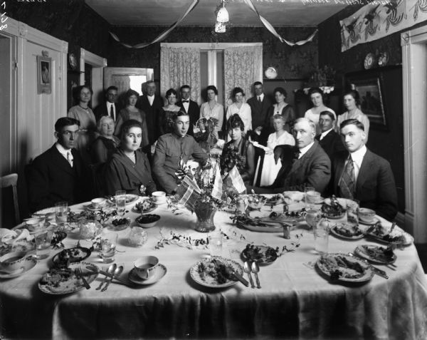 Some twenty members of the Harold Stafford wedding party and guests, seated and standing behind a table set for a meal, with food on many of the plates. The groom is in military uniform. There is a centerpiece on the table of flowers and small flags. Streamers hang from the ceiling.