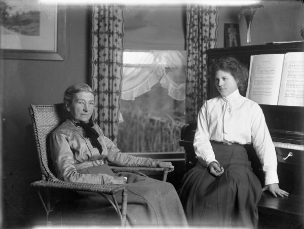 Mrs. Pierce sits in a wicker armchair facing the camera while her granddaughter Margery Bish sits facing her on a stool in front of a piano. Behind them is a window with open curtains. Sheet music open on the piano.