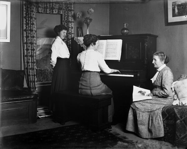 Mary Pierce Bish plays an upright piano while her daughter Margaret Bish, standing to her left, and her mother Mrs. Pierce, seated to her right, look on.