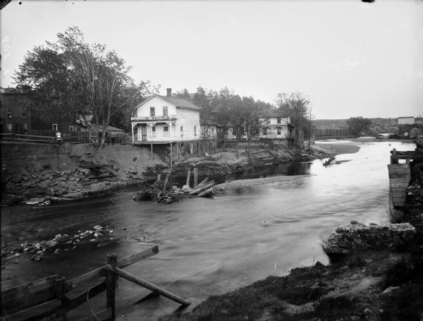 View across river of several houses and other buildings standing at the edge of the Chippewa River. One building has stilts supporting the back porch over the steep river bank. There is debris in the river, and the shoreline shows damage from flooding.