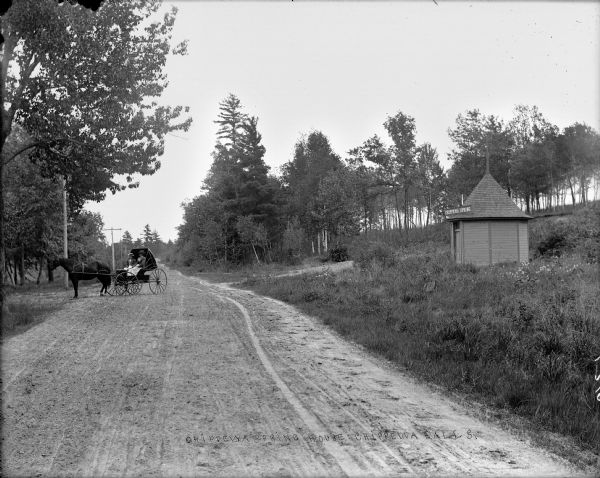 The Chippewa Spring House, a small round building with a conical roof with a sign over the door reading "Chippewa Spring," sitting off to the side of a dirt road in tall grass. Two women wearing straw hats sit in a buggy pulled by two horses are parked on the road.