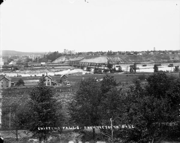 Elevated view from south side of the Chippewa River, with a bridge crossing it and buildings on both sides of the river. A bluff rises on the far side of the river and trees are in the foreground.