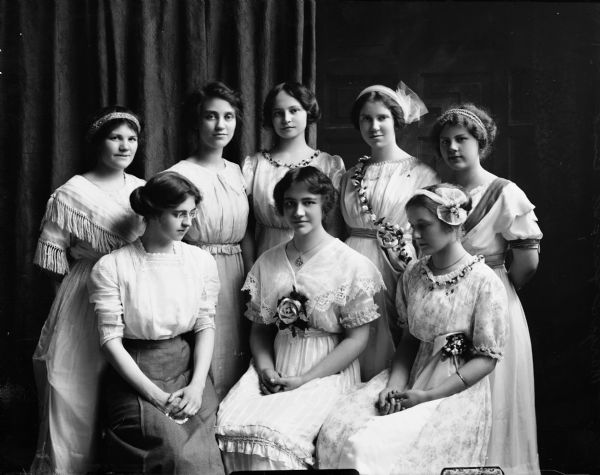 Margaret Bish, the daughter of the photographer, sits in the center front row of two rows of young women, all dressed in white dresses, many decorated with flowers.