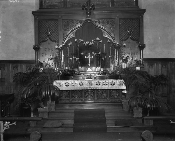 The raised altar of the Episcopal Church, with potted palms sitting on the steps. Lilies in vases and candles are displayed on the altar. Elaborate woodwork decorates the altar and the wall behind it.