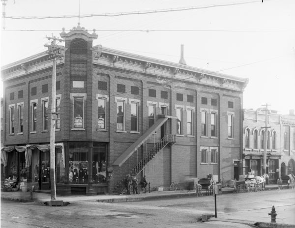 View form across street of the French Lumber Company, a two-story brick building with the Chicago Dental Parlors occupying the second floor, on a street corner. Three men stand in front of a fire escape leading to the second floor. Several buggies, one with horses hitched to it, stand on the street. There is a utility pole in front of the building.