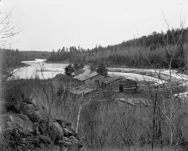 View from hill looking down on a group of log cabins inhabited by "dam tenders" on the shore of the icebound Chippewa River. A logging dam in is in the right forefront. The shore of the river is heavily wooded.