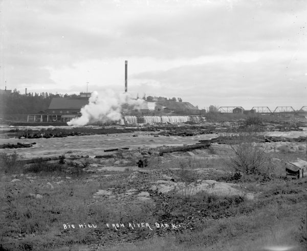 View from shoreline of lumber mill on the bank of the Chippewa River, with steam emitting from the smokestacks. A bridge over the river is in the right background.