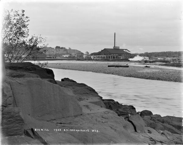 Elevated view from steep shoreline opposite a lumber mill on the far shore of the Chippewa River, with a bluff rising above it to the right. Boulders are on the shoreline in the foreground.